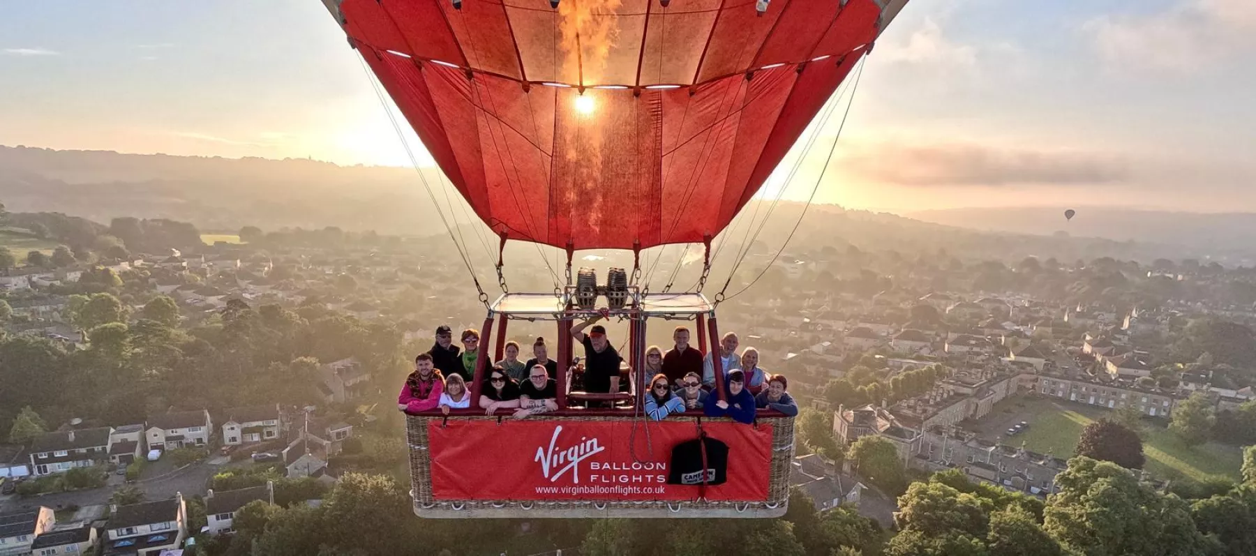 A red Virgin hot air balloon rising into the sky over a sun-soaked misty skyline of Bath in Somerset