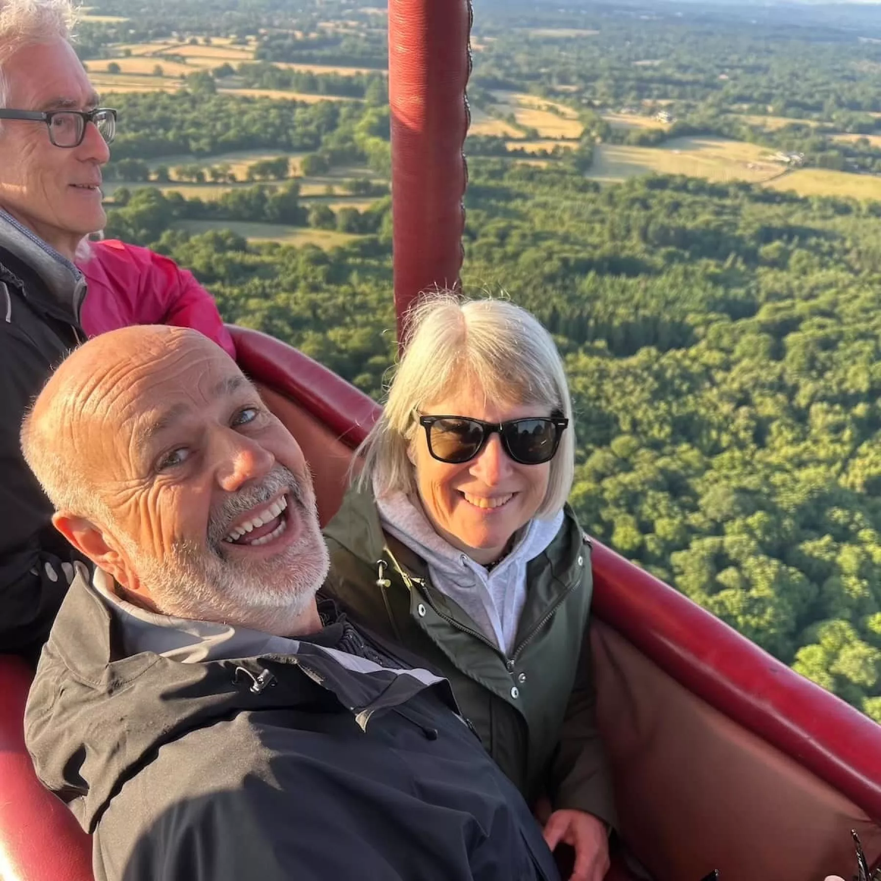 A couple laughing together in a hot air balloon basket, with views of fields and woodland in the background